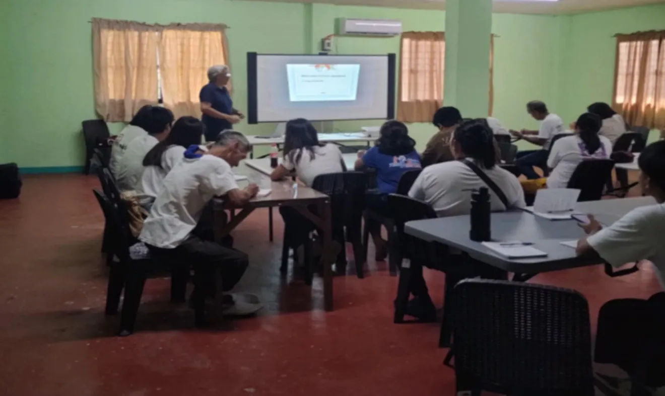 Adult learners seated at tables in a bright classroom, watching a presentation on a screen.